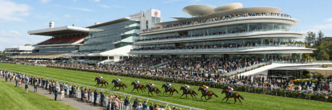 A horse race on a green turf track with several horses and jockeys racing past a large multi-tiered white stadium filled with spectators at a sunny day festival.
