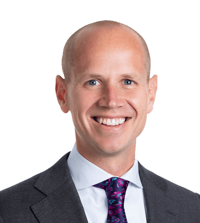 Portrait of a smiling bald man in a dark suit, white shirt, and purple patterned tie, facing the camera.