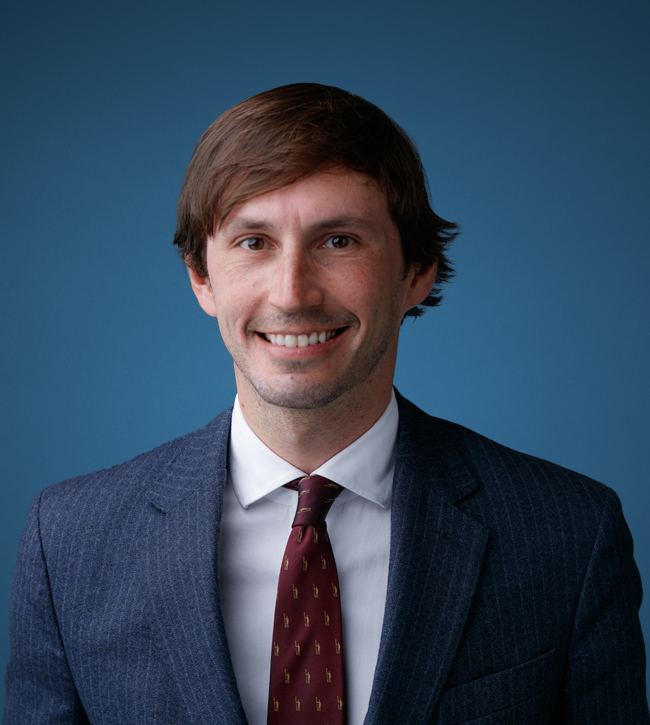 daniel-romagnolo_ Head-and-shoulders portrait of a smiling man in a blue pinstripe suit and burgundy tie against a blue background
